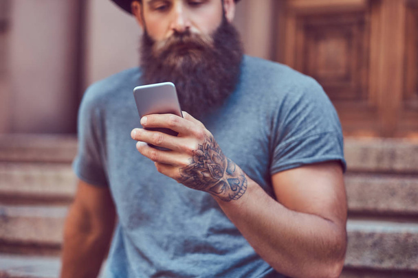 Handsome hipster male with a stylish beard and a tattoo on his arms dressed in casual clothes and hat using smartphone while sitting on steps. - Fotó, kép