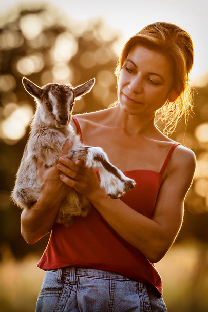 Beautiful woman with a small and nice goatling on her hands in a sunset background - Photo, Image