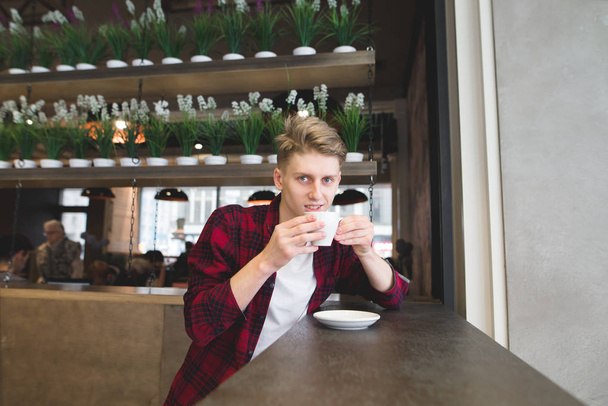 A positive young man drinks coffee in a cozy cafe and looks at the camera. Lunch in a cozy cafe - Photo, Image