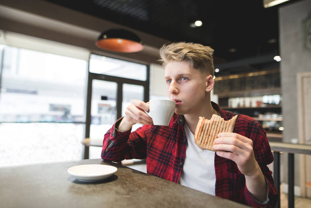 Un joven come un sándwich y bebe café en un acogedor café. Los platos del estudiante en un café y mira cuidadosamente en la ventana
 - Foto, imagen