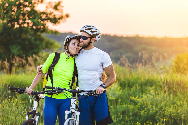 Een man en een vrouw fietsen staan samen in een park in de buurt van hun fiets in het voorjaar bij zonsondergang. De man omarmd de vrouw. - Foto, afbeelding