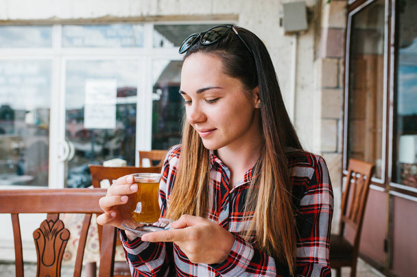 Красива молода дівчина з сонцезахисними окулярами на голові сидить за столом у відкритому кафе та п'є чай зі скляної склянки
 - Фото, зображення