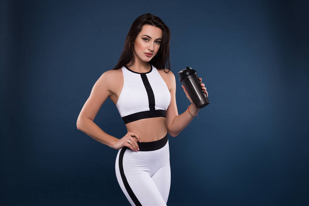 Stay strong and fit. Attractive young sporty woman with protein shake bottle on dark blue background pouncing for the picture in studio. - Zdjęcie, obraz