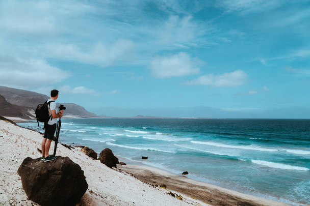 Photographer with stative and camera staying on the rock and enjoying coastal landscape of sand dunes, volcanic cliffs and ocean waves. Baia Das Gatas, near Calhau, Sao Vicente Island Cape Verde - Zdjęcie, obraz
