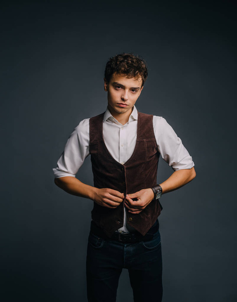 A young, handsome guy posing on a gray studio background. The man is stylish and elegantly dressed in jeans, a shirt and a brown vest. - Valokuva, kuva