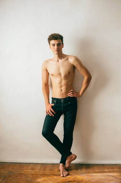 A young, handsome guy posing with a gnawed torso, demonstrating a sports body, muscles, relief, a pumped-up body. Background white wall. Shooting with natural light, minimal, natural retouching - Foto, Imagen
