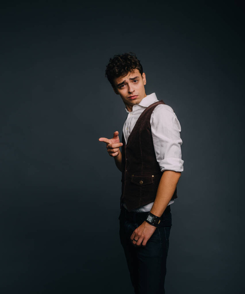 A young, handsome guy posing on a gray studio background. The man is stylish dressed. The guy calls attention and points a finger at the object causing astonishment and disgust - Foto, Imagem