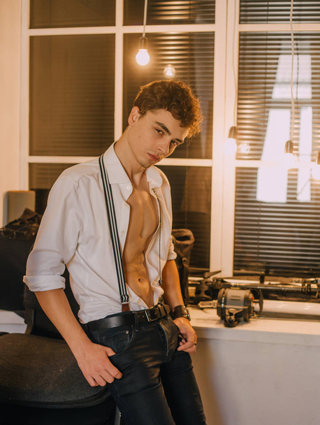 A young, handsome guy posing with an unbuttoned, white shirt, with braces in retro style, demonstrates the athletic body. The background is a large window and a lamp. minimal, natural retouching - Фото, зображення