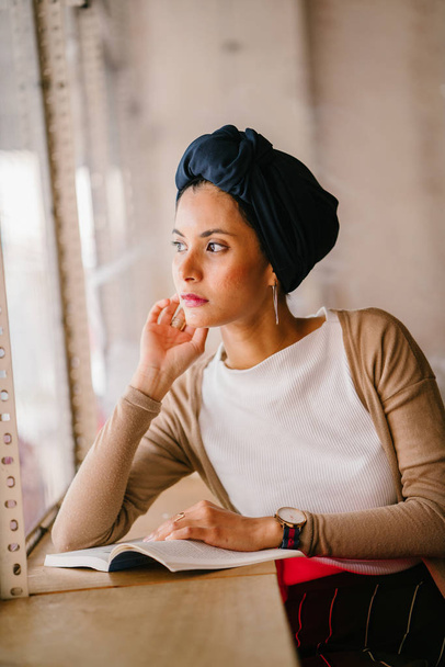 young, attractive Muslim woman (Arab, Malay, Asian) reads a book by the window of a cafe in the day. She is wearing a turban (headscarf, hijabi) and is elegantly dressed in earthy tones. - 写真・画像