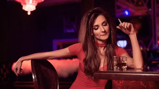 Portrét nádherná krása bruneta dívka v červených šatech sedí u baru se sklenkou whisky a cigaret v luxusní interiér - Záběry, video