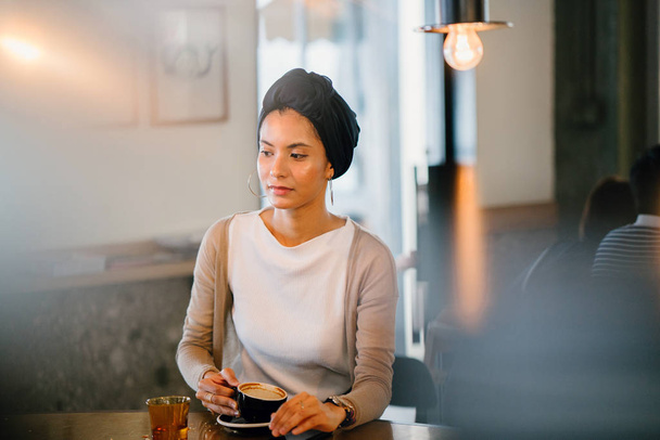 young, attractive Muslim woman (Arab, Malay, Asian)with coffee in  cafe in the day. She is wearing a turban (headscarf, hijabi) and is elegantly dressed in earthy tones. - Foto, immagini