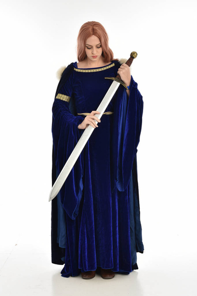 retrato de longitud completa de la mujer con el pelo largo, con un vestido medieval de terciopelo azul y capa de piel. pose de pie sosteniendo una espada larga, aislada sobre fondo blanco
. - Foto, imagen