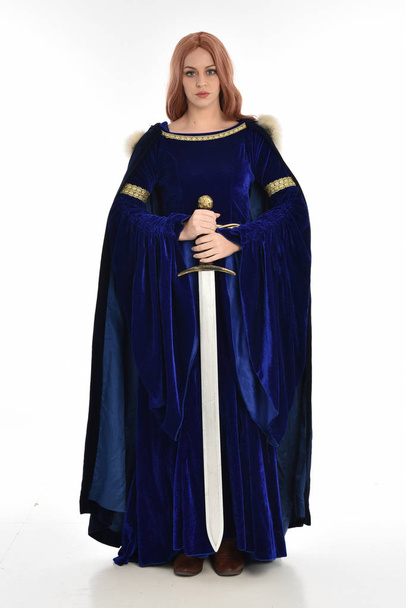 retrato de comprimento total da mulher com cabelos longos, vestindo um vestido medieval de veludo azul e capa de pele. postura de pé segurando uma espada longa, isolada no fundo branco
. - Foto, Imagem