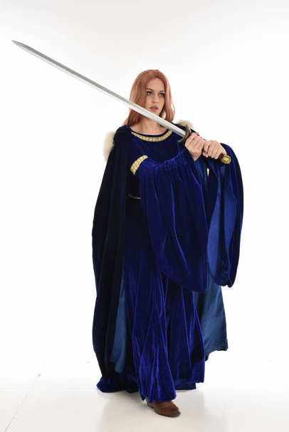 Pełna długość Portret kobiety z długimi włosami, noszenie blue velvet średniowiecznych suknia i futro płaszcz. Stojący poza trzymając długi miecz, na białym tle. - Zdjęcie, obraz