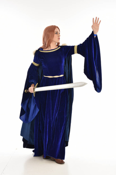 portrait complet de femme aux cheveux longs, vêtue d'une robe médiévale en velours bleu et d'un manteau de fourrure. pose debout tenant une longue épée, isolé sur fond blanc
. - Photo, image