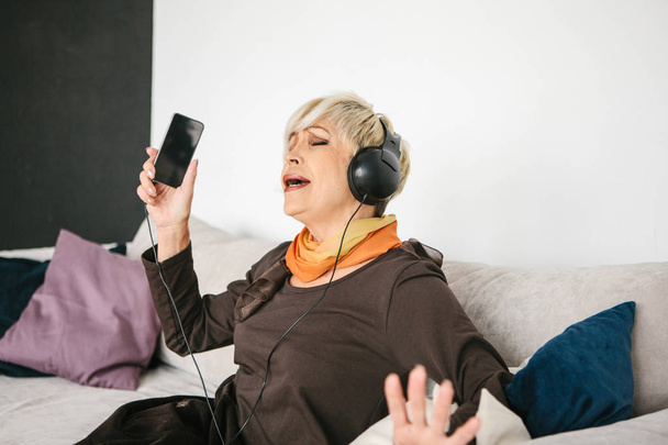 Μια θετική ηλικιωμένη γυναίκα ακούει μουσική και τραγουδάει. Η παλαιότερη γενιά και οι νέες τεχνολογίες. - Φωτογραφία, εικόνα