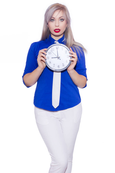 Studio business portrait of a beautiful young blonde woman in a dark blue shirt, white trousers and a white tie. The girl is holding a clock. Isolated. Copy space - Foto, Imagem