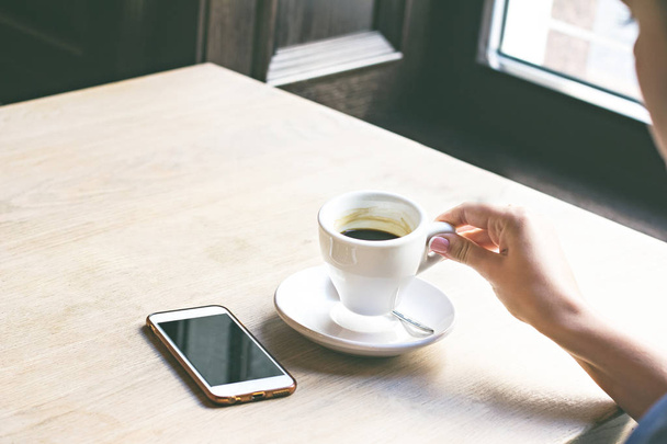 Nahaufnahme des Mobiltelefons in den Händen der Frau, sitzend mit einer Tasse Kaffee.  - Foto, Bild