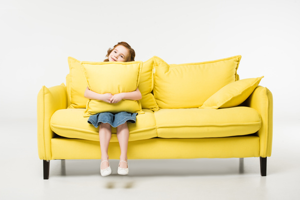 Kleines Kind mit Kissen in den Händen sitzt isoliert auf weißer Couch - Foto, Bild