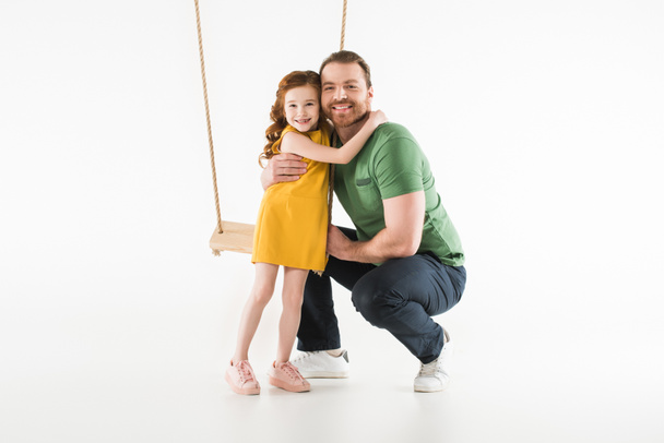 Κόρη στέκεται κοντά σε swing και αγκαλιάζει τον πατέρα που απομονώνονται σε λευκό - Φωτογραφία, εικόνα