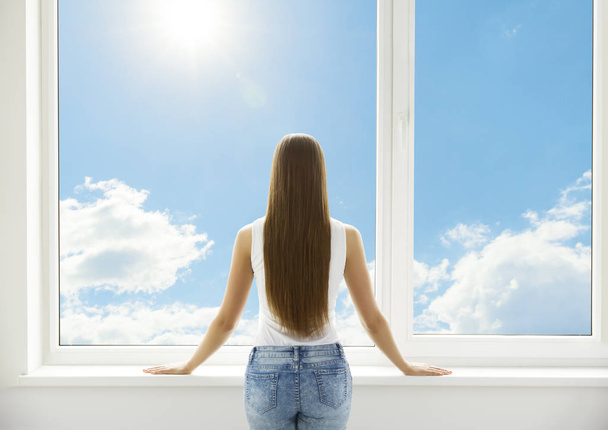 Окно и женщина, вид сзади на молодую девушку, стоящую в интерьере Белого дома, человек смотрит через окно ПВХ
 - Фото, изображение