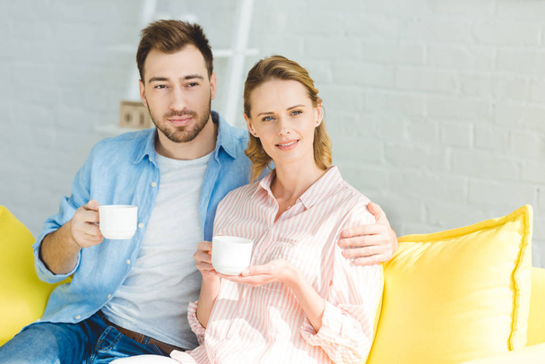 Nuori pari istuu sohvalla kupit teetä käsissä
 - Valokuva, kuva
