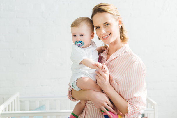 Smiling mother holding infant daughter with baby dummy - Fotografie, Obrázek