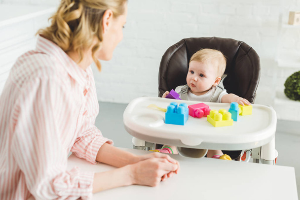 Madre guardando la figlia neonata seduta sulla sedia del bambino con blocchi di plastica
 - Foto, immagini