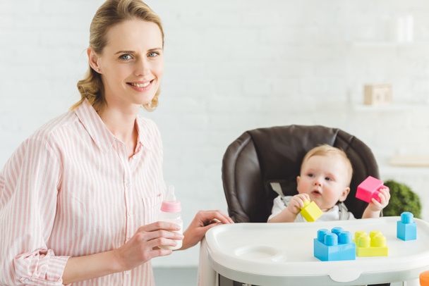 Young mother with feeding bottle and infant daughter sitting in baby chair with plastic blocks - Foto, imagen