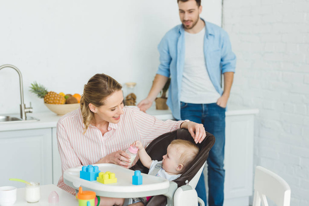 Mother giving feeding bottle to infant daughter in baby chair while father standing behind - Photo, Image