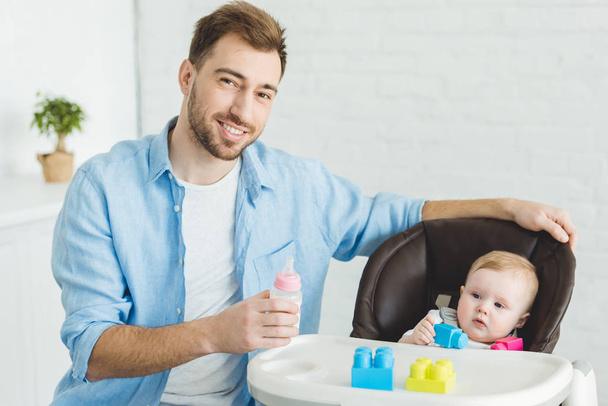 Uśmiechnięty ojciec z butelką i córeczka w krzesełko dla dziecka z plastikowych klocków - Zdjęcie, obraz