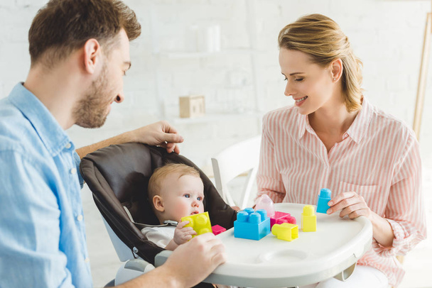Smiling parents and infant daughter in baby chair with plastic blocks - Foto, imagen