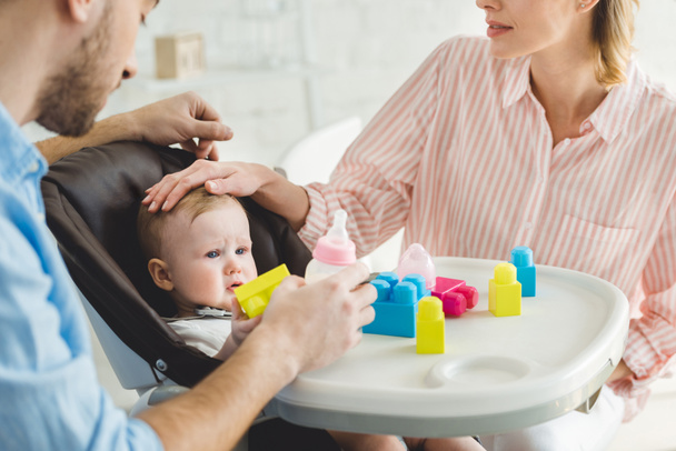 Vista recortada de los padres con biberón e hija lactante en silla de bebé con bloques de plástico
 - Foto, imagen