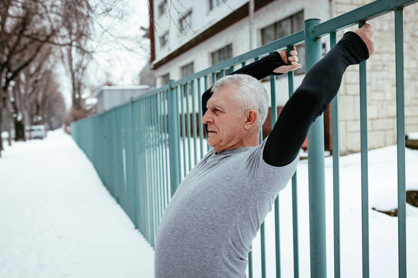 Активний старший чоловік розтягується і виконує вправи, використовуючи паркан під час зимових тренувань зовні в
. - Фото, зображення