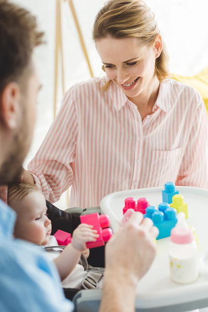 Ouders met dochtertje zit in kinderstoel met kunststof blokken en voedingsfles - Foto, afbeelding