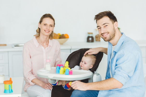 Young parents with infant daughter sitting in baby chair with plastic blocks and feeding bottle - Photo, Image