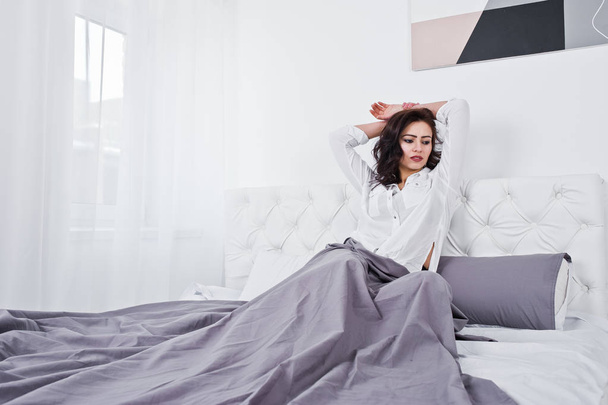 Studio portrait of brunette girl in white blouse lying on bed. - Фото, изображение
