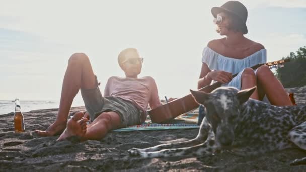 Young couple having fun on the beach. Weekend on the island. Young couple singing with a guitar on the beach their funny dog. Picnic on the seaside - Materiał filmowy, wideo