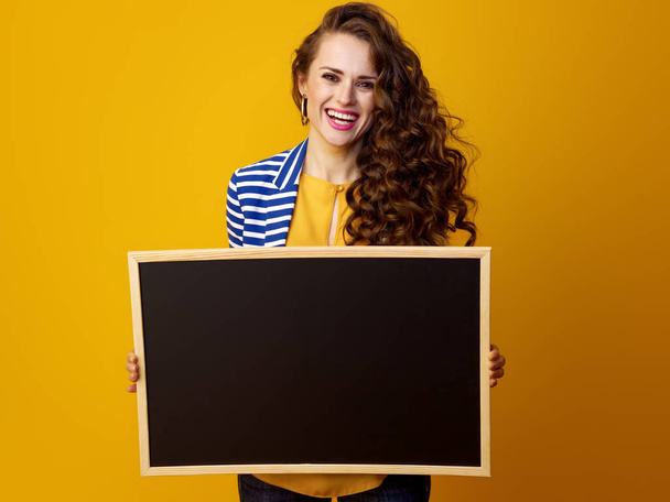 smiling stylish woman with long wavy brunette hair against yellow background showing blank board - Фото, изображение