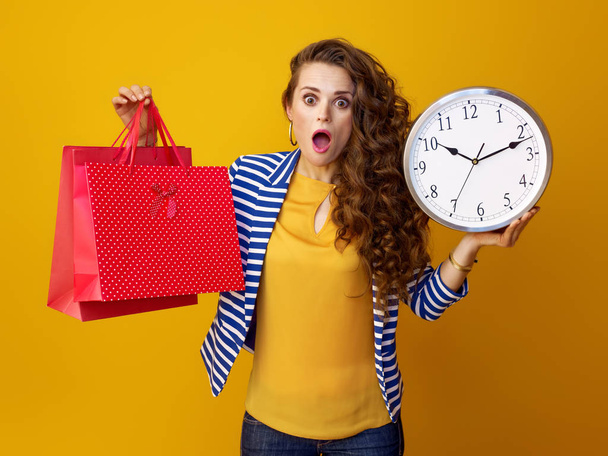 surprised trendy woman with long wavy brunette hair against yellow background showing clock and red shopping bags - Photo, image