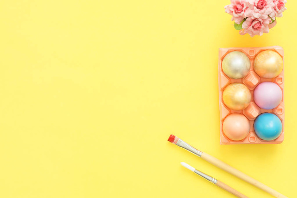 Flat lay top view colorful easter egg painted in pastel colors composition and spring flowers with paint brush on yellow pastel color background. Easter day background top view with copy space. - Photo, Image