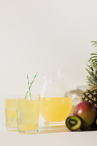 Nahaufnahme von Gläsern und Glaskannen mit Saft und frischen tropischen Früchten auf grau  - Foto, Bild