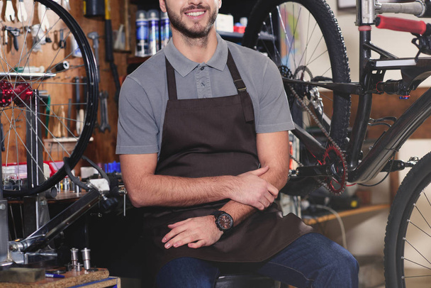 Schnappschuss eines lächelnden jungen Mannes in Aron, der in der Nähe von Fahrrädern in der Werkstatt sitzt - Foto, Bild