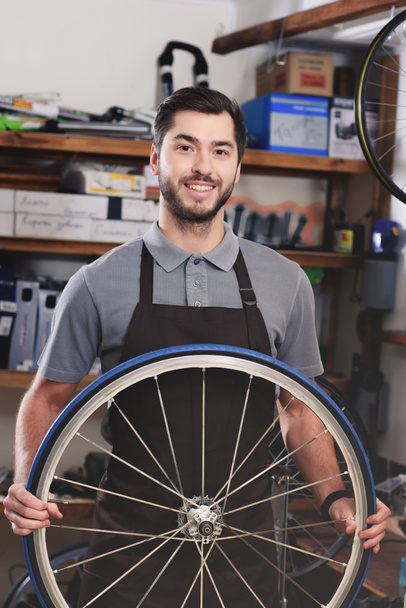 jovem trabalhador bonito no avental segurando roda de bicicleta e sorrindo para a câmera na oficina
 - Foto, Imagem