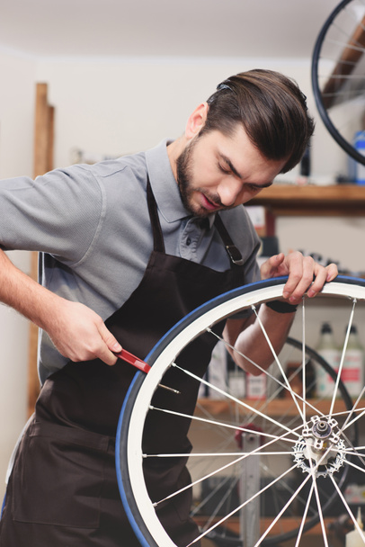 handsome young worker in apron fixing bicycle wheel in workshop   - Photo, Image