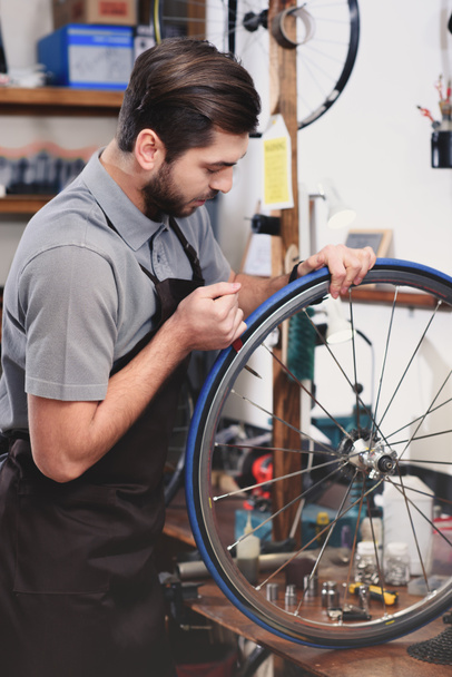 young mechanic in apron repairing bicycle wheel in workshop  - Fotó, kép