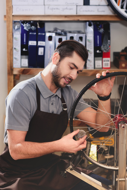 smiling young man in apron fixing bicycle wheel in workshop  - Фото, изображение