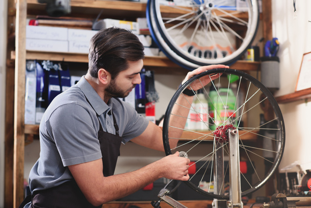 young man in apron repairing bicycle wheel in workshop  - Foto, Imagen