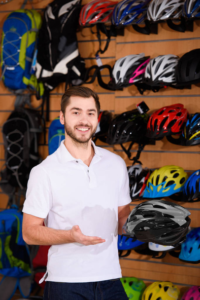 handsome young seller holding bicycle helmet and smiling at camera in bike shop - Φωτογραφία, εικόνα