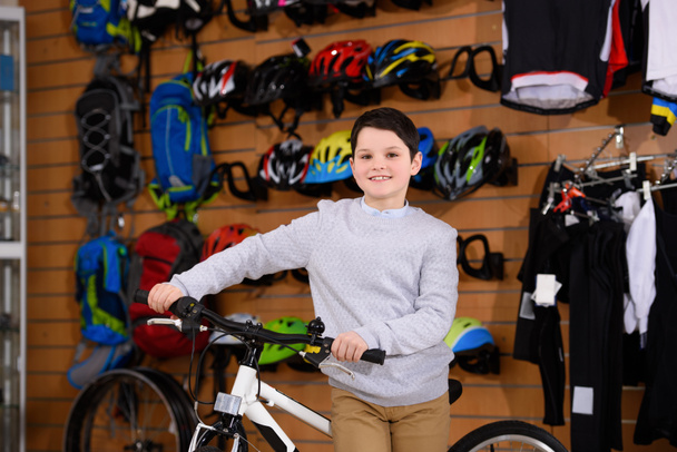 cute little boy standing with bicycle and smiling at camera in bike shop  - Photo, Image